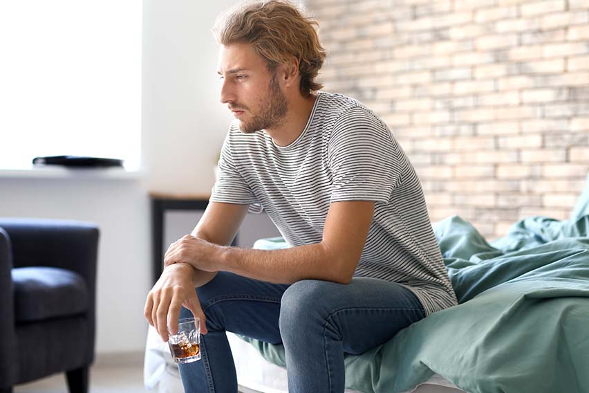 man sitting on the end of his bed with a glass of liquor experiencing delirium tremens